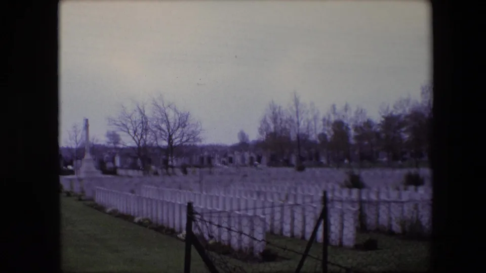 1967 Calais 8mm frame showing rows of white military cemetery gravestones, a grassy path, bare trees, and a distant stone memorial under gray sky.