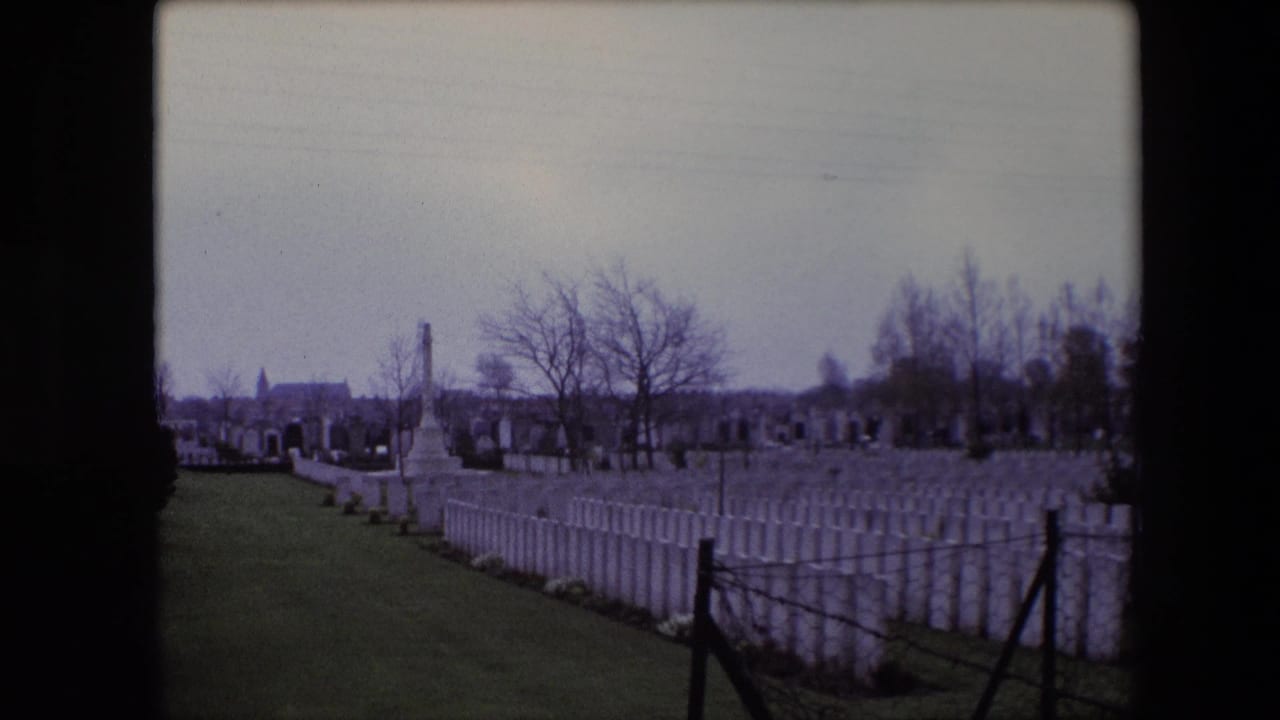 Military cemetery rows with a stone memorial, bare trees, and distant buildings beyond the graves.