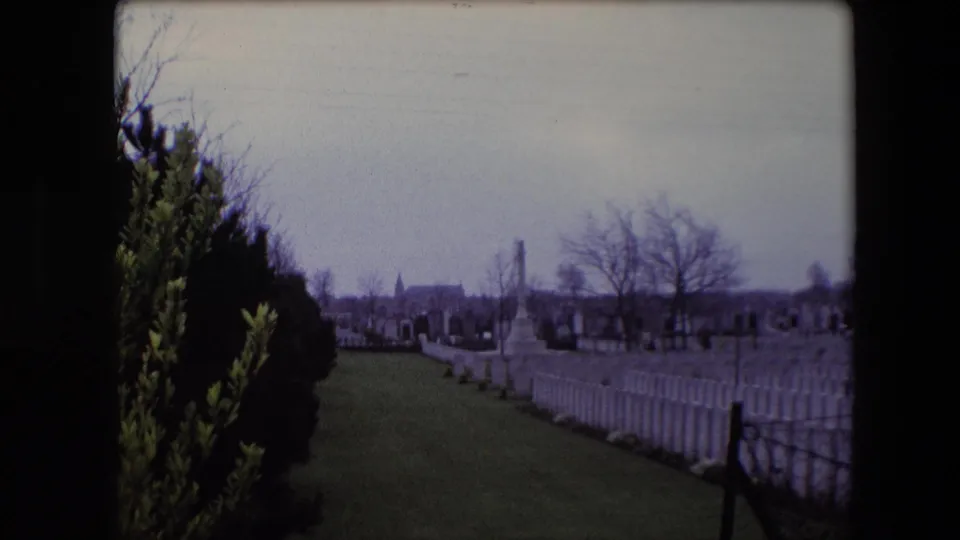 1967 Calais 8mm frame looking past dark shrubs toward white military graves, open grass, a low fence, bare trees, and a distant memorial.