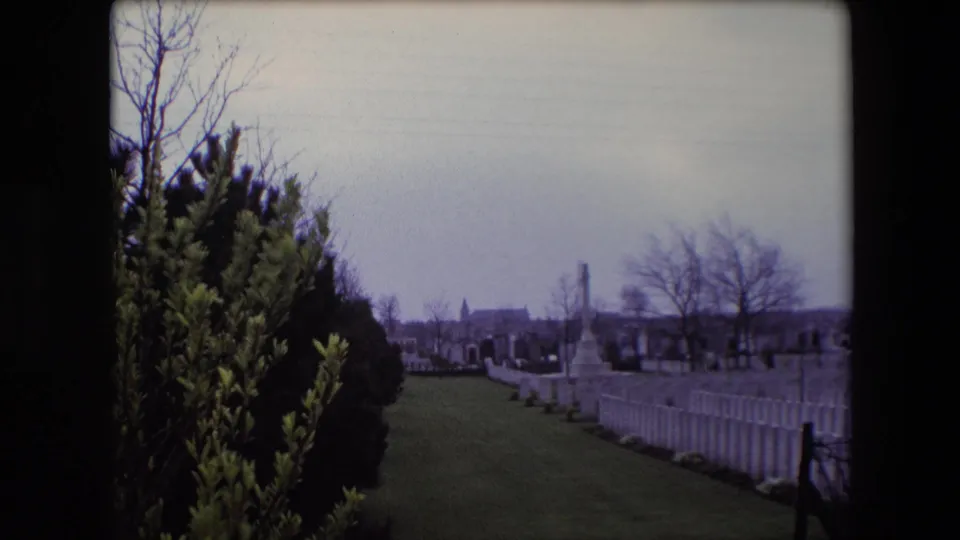 1967 Calais cemetery frame with foreground shrubs, rows of white military gravestones, trimmed grass, bare trees, and a tall memorial in the distance.