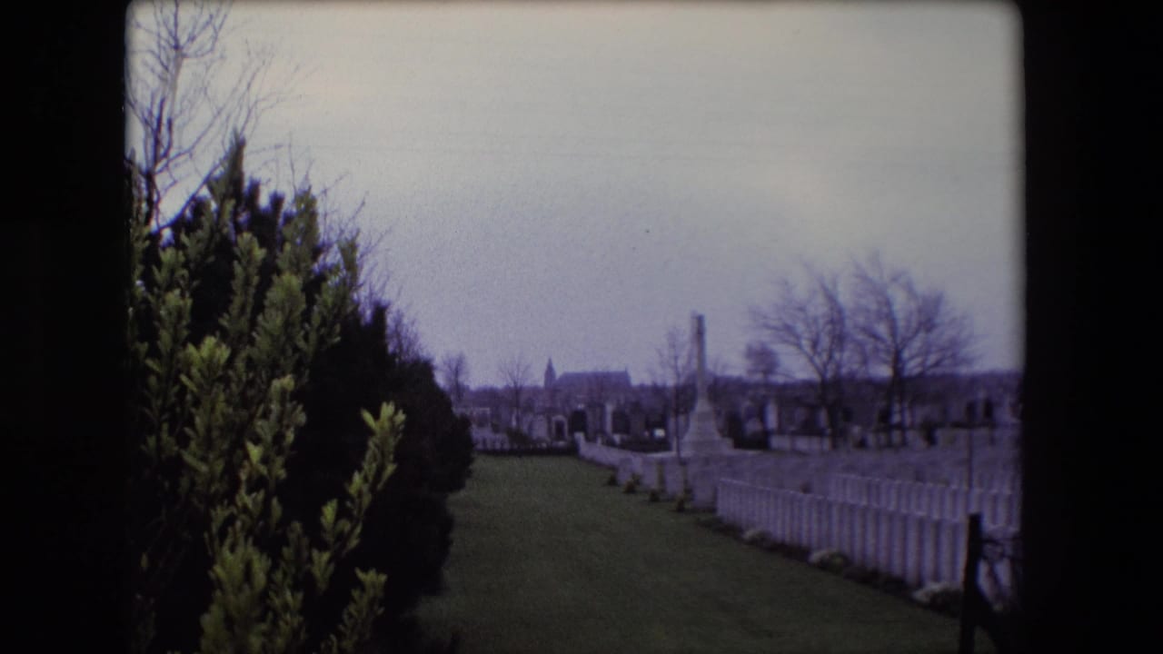 1967 Calais home-movie view of a cemetery lawn with white headstones, bare trees, shrubs, and a distant church-like building.