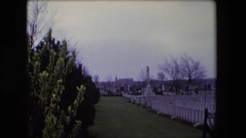 1967 Calais home-movie view of a cemetery lawn with white headstones, bare trees, shrubs, and a distant church-like building.