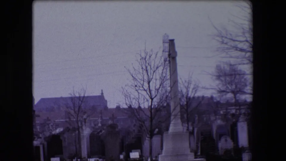 1967 Calais footage frames a tall cross-shaped war memorial within a cemetery, with headstones, bare trees, and a church roofline behind it.