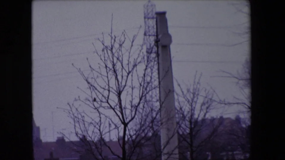 Close 1967 Calais cemetery view of a tall stone memorial or cross, partially hidden by bare branches, with distant buildings beyond.