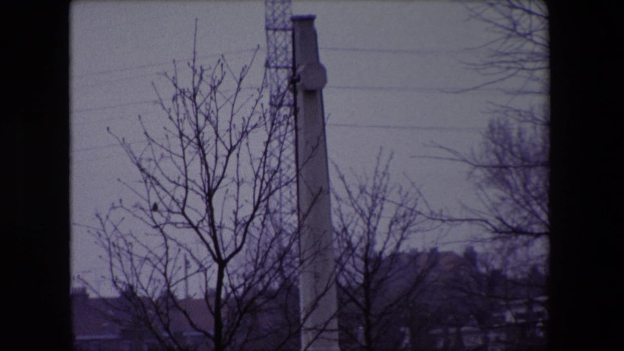 1967 Calais cemetery footage showing bare branches, a tall stone memorial, distant rooftops, and a tower or mast under a gray sky.