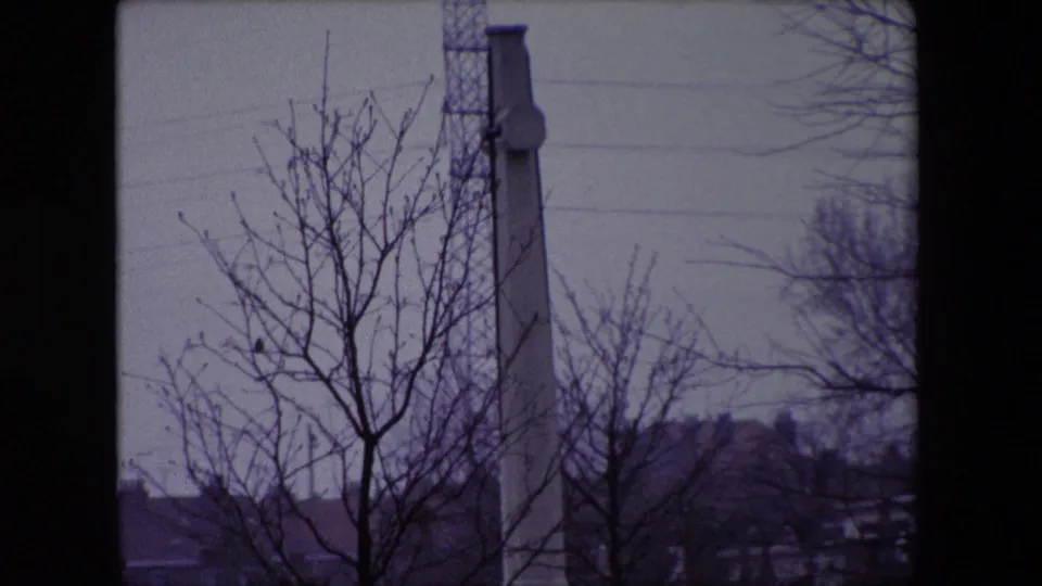1967 Calais cemetery footage showing bare branches, a tall stone memorial, distant rooftops, and a tower or mast under a gray sky.