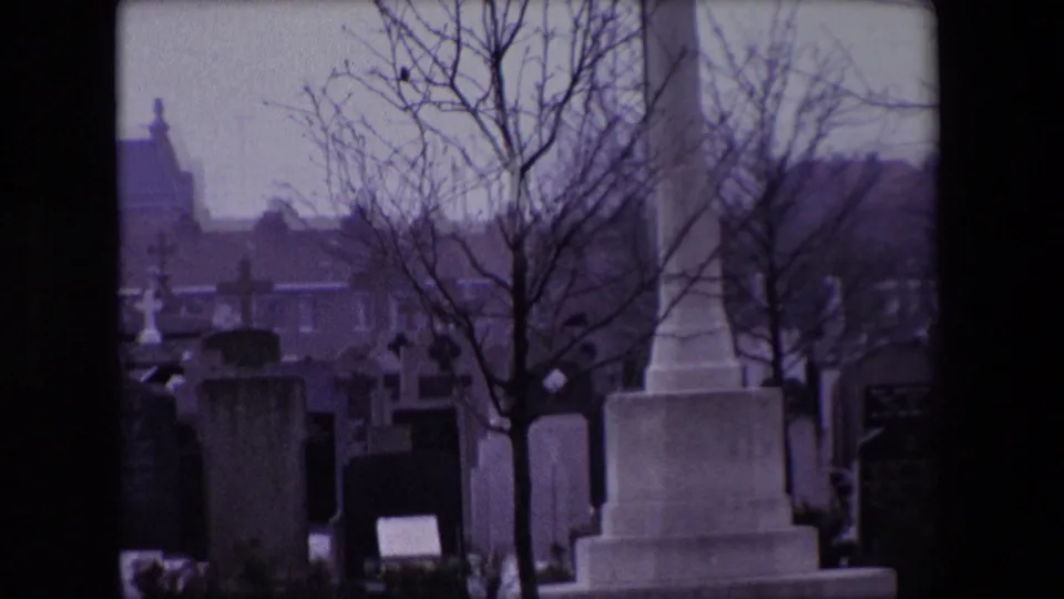 Cemetery view in Calais, France, with stone monuments, headstones, bare trees, and distant buildings visible through a dark film gate.