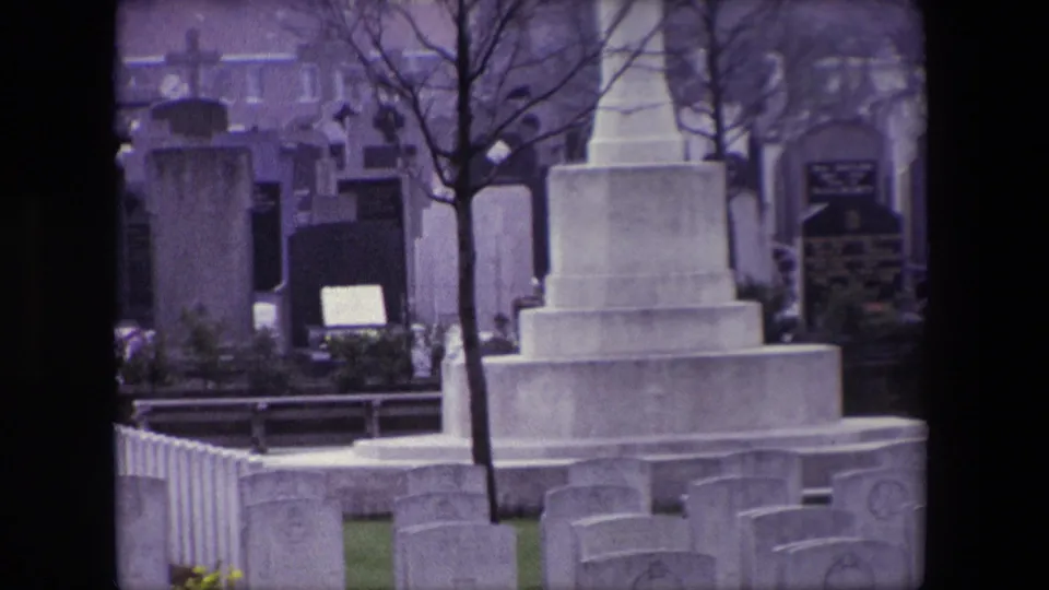 Closer 1967 Calais cemetery view of a stepped stone memorial, surrounding grave markers, bare branches, and buildings beyond the cemetery.
