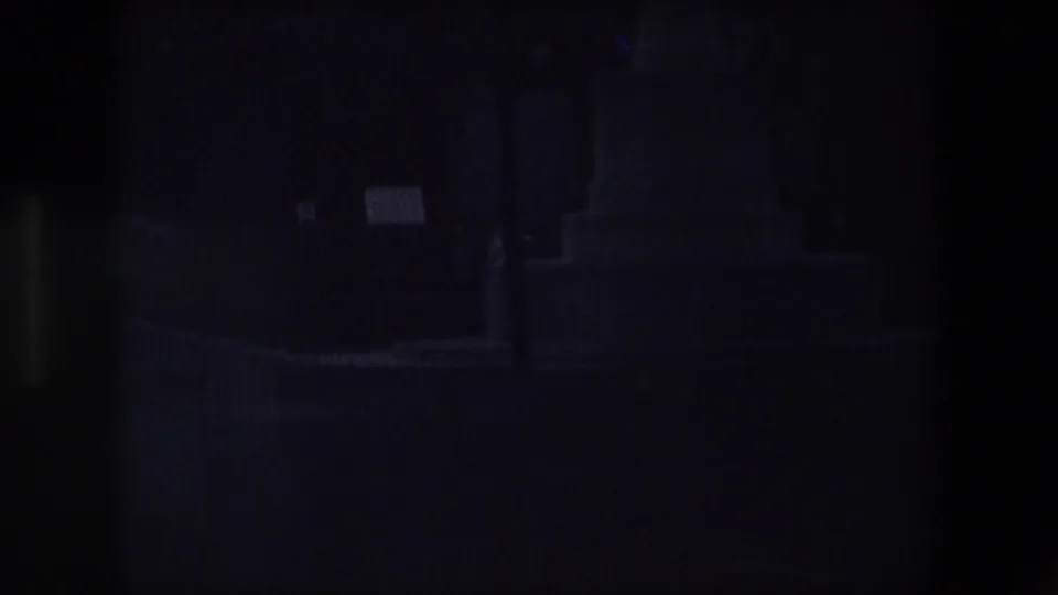 Dark underexposed 8mm frame from the Calais cemetery scene, with faint outlines of monuments, trees, and a small pale marker still visible.