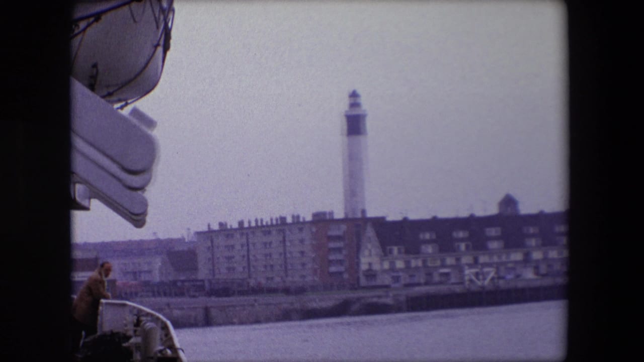 1967 Calais home-movie frame from a vessel, showing an adult passenger at the deck edge with a white lighthouse, seawall, and waterfront buildings in the distance.