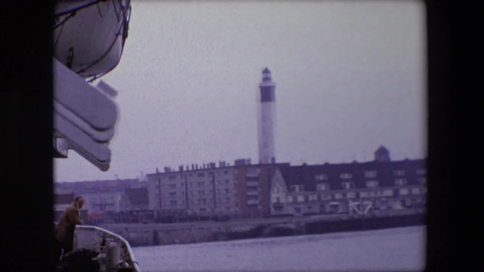 1967 Calais home-movie frame from a vessel, showing an adult passenger at the deck edge with a white lighthouse, seawall, and waterfront buildings in the distance.