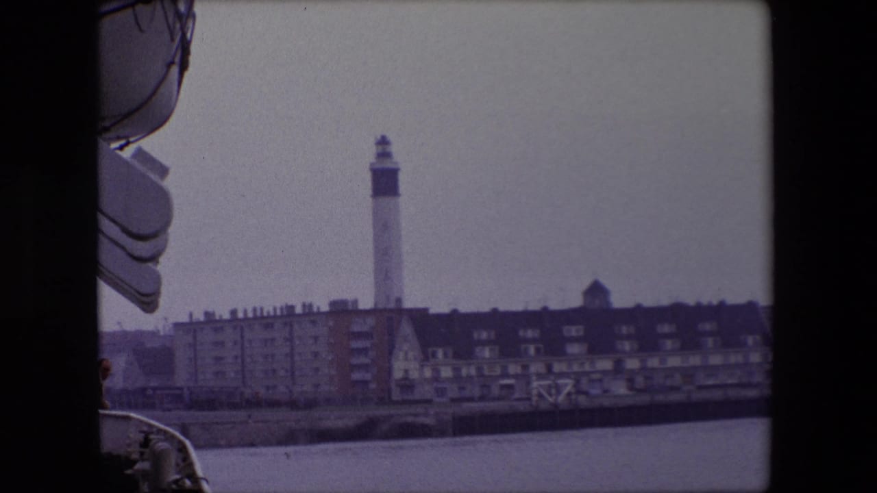 Shipboard view of a lighthouse behind waterfront buildings and a seawall.