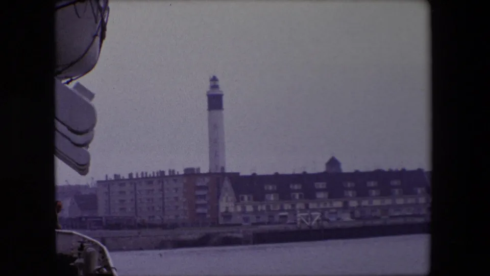 1967 Calais harbor view from a moving vessel, with ship structure at the left edge and a white lighthouse rising above waterfront buildings under an overcast sky.