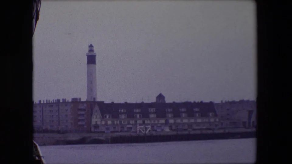 1967 Calais waterfront frame showing the lighthouse behind a row of buildings, with harbor water in the foreground and a small portion of ship structure at the lower left.