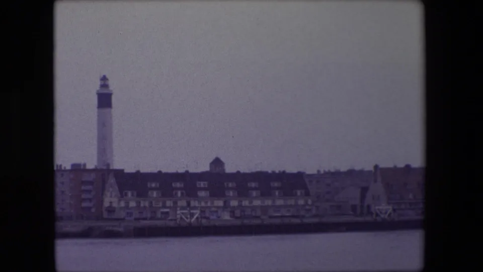 1967 Calais harbor frame with the white lighthouse and low waterfront buildings across the water, recorded in muted 8mm color from a ship.