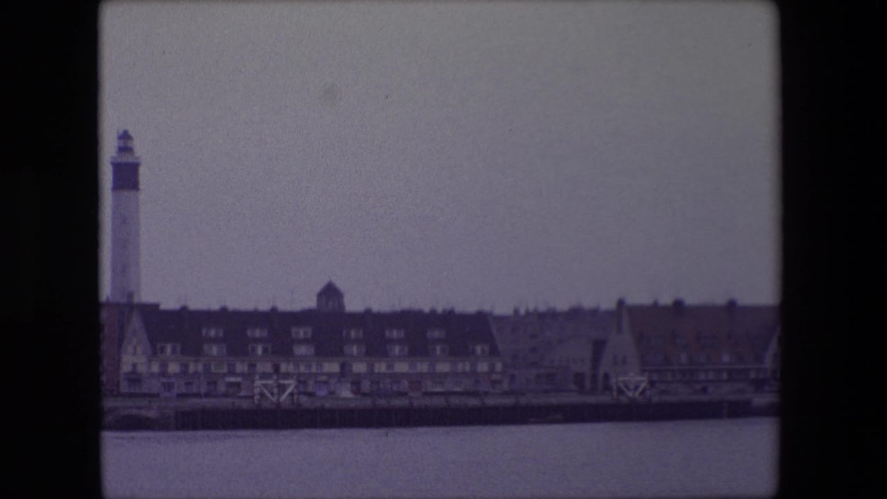 1967 Calais, France waterfront view with still water, dark-roofed buildings, gabled facades and a tall lighthouse-like harbor tower at the left.
