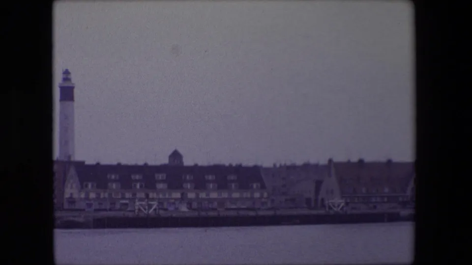 1967 Calais, France waterfront view with still water, dark-roofed buildings, gabled facades and a tall lighthouse-like harbor tower at the left.