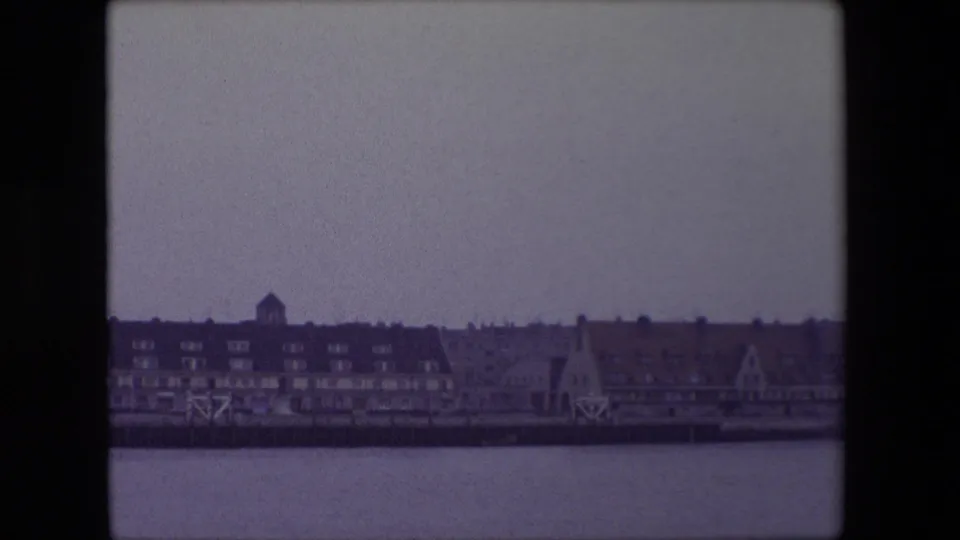 1967 Calais riverfront or harborfront buildings seen from across the water, with a dark roofline, small tower form and pale winter sky.