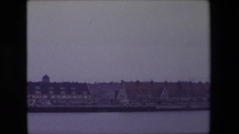 1967 Calais waterfront view after the pan, showing a row of low buildings, steep rooflines, quay structures and still water in muted winter light.