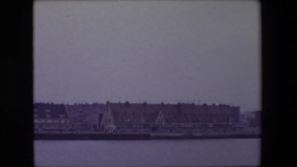 1967 Calais harborfront or riverfront scene with a continuing line of masonry buildings, dark roofs, a quay edge and calm water under an overcast sky.
