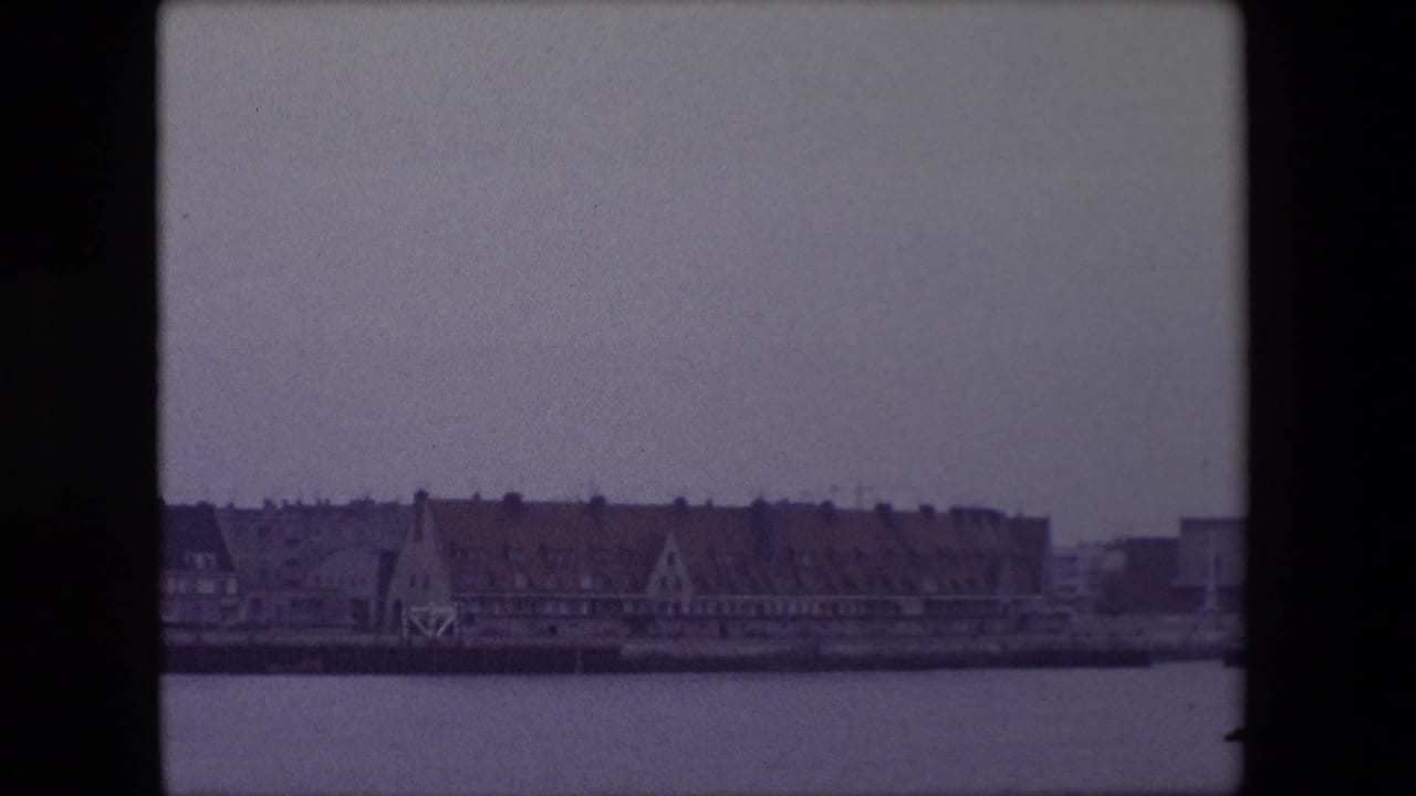 1967 Calais waterfront view across harbor water, with red-tiled buildings and a distant steeple under a muted overcast sky.