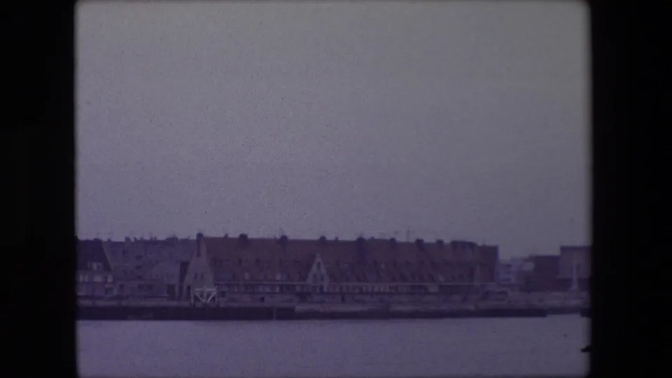 1967 Calais waterfront view across harbor water, with red-tiled buildings and a distant steeple under a muted overcast sky.