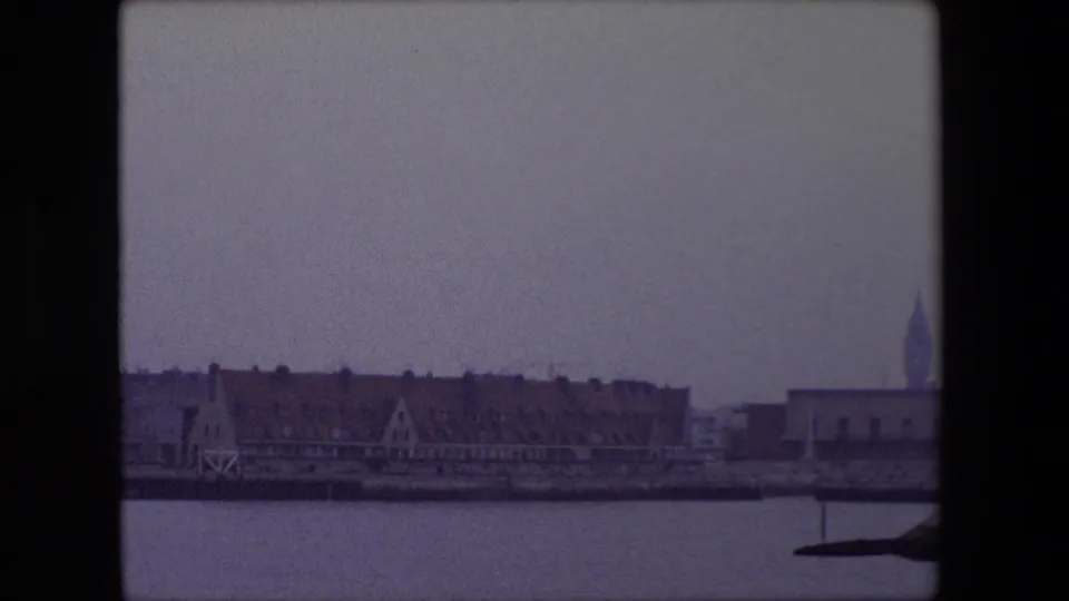 Calais, France waterfront in 1967, showing low harbor buildings, red roofs, calm water, and a distant church steeple near the right side of the frame.