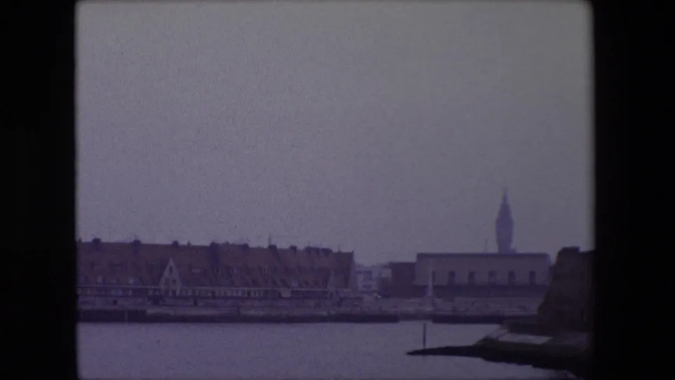 1967 Calais harbor panorama with red-roofed waterfront buildings, a distant steeple, and a dark stone foreground structure entering at lower right.