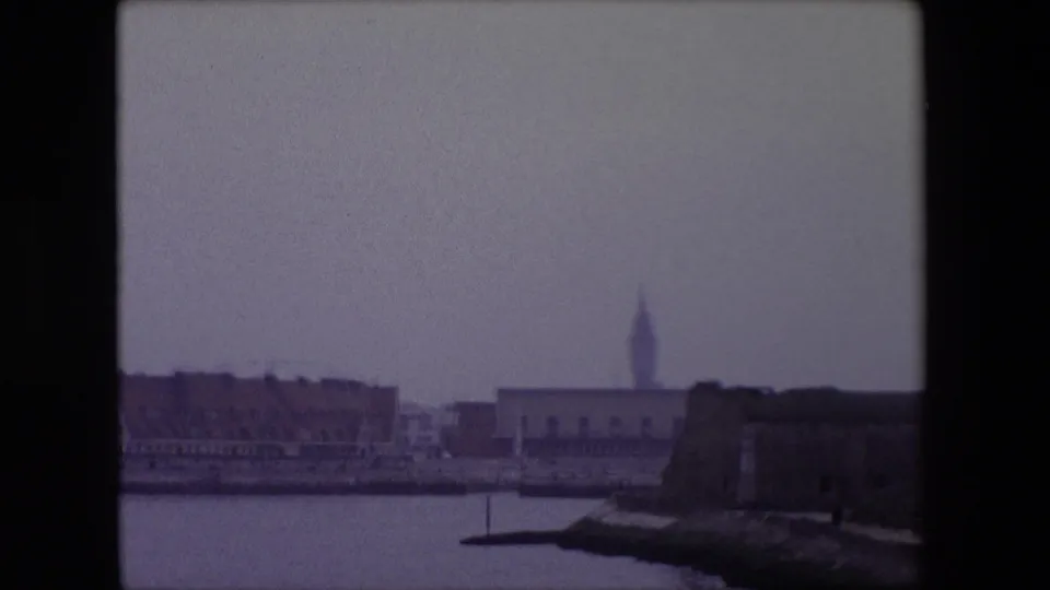 End of the 1967 Calais waterfront pan, with harbor water, low town buildings, a distant church steeple, and a dark stone fortification-like foreground form.