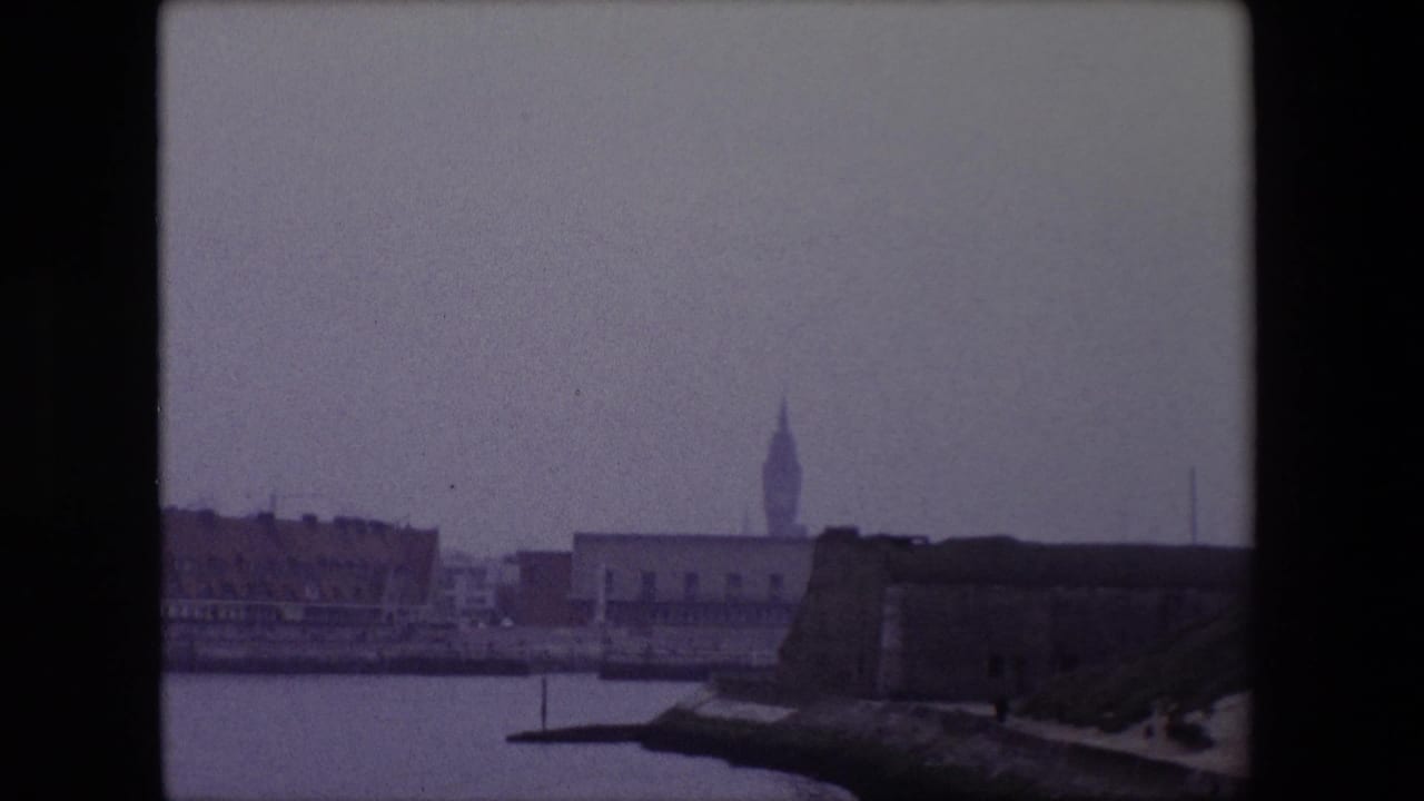 1967 Calais home-movie frame showing harbor water, a low fortified waterfront structure, distant city buildings, and a pointed landmark tower in soft 8mm color.