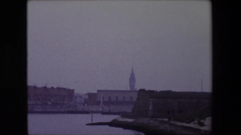 1967 Calais home-movie frame showing harbor water, a low fortified waterfront structure, distant city buildings, and a pointed landmark tower in soft 8mm color.