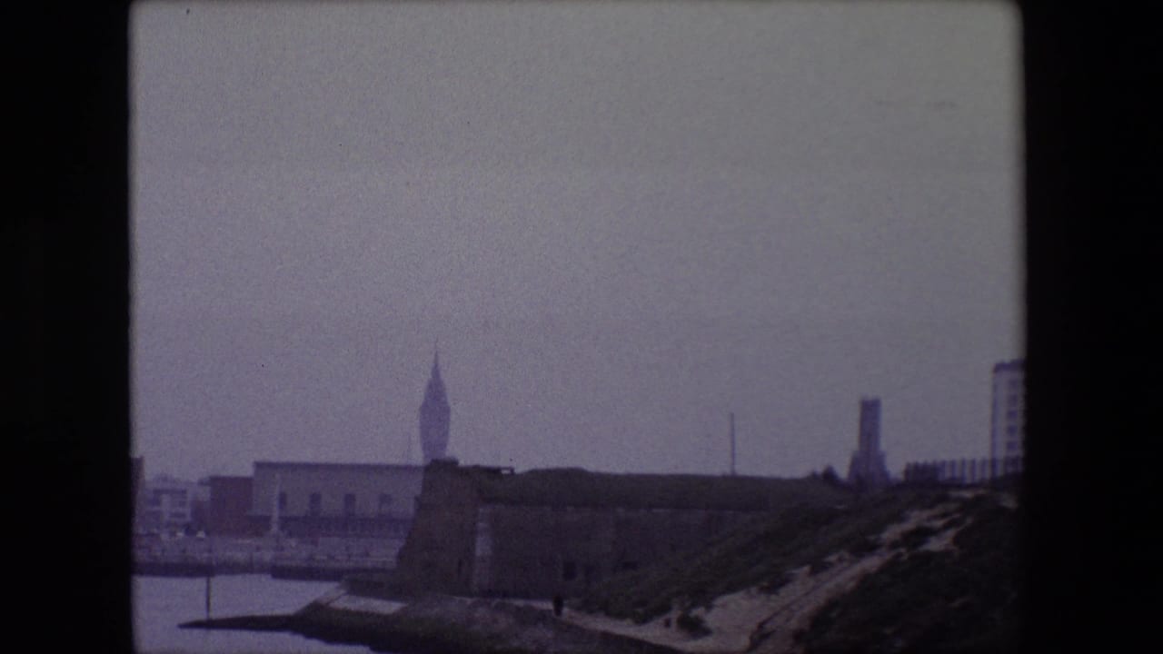 1967 Calais frame from a panning 8mm view, with the pointed tower, harbor edge, old fortification, sloped embankment, and distant buildings under overcast haze.