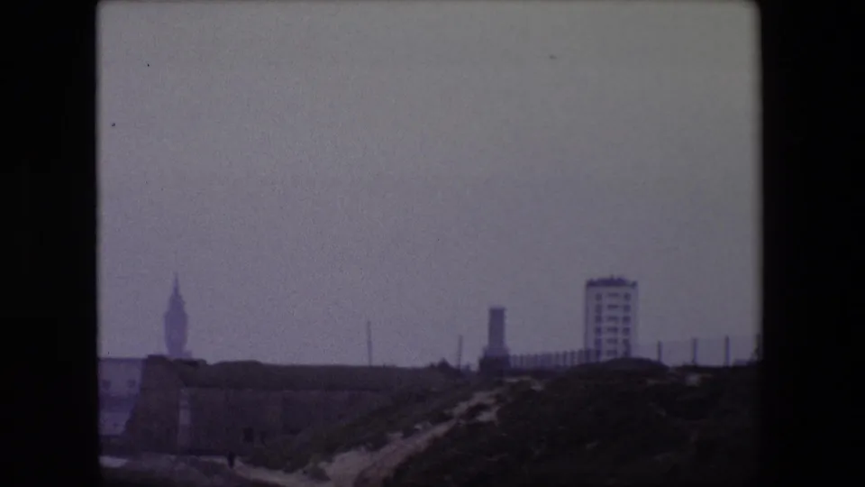 1967 Calais 8mm frame showing the pan moving toward a fenced embankment and modern rectangular building, with the pointed tower receding at the left edge.