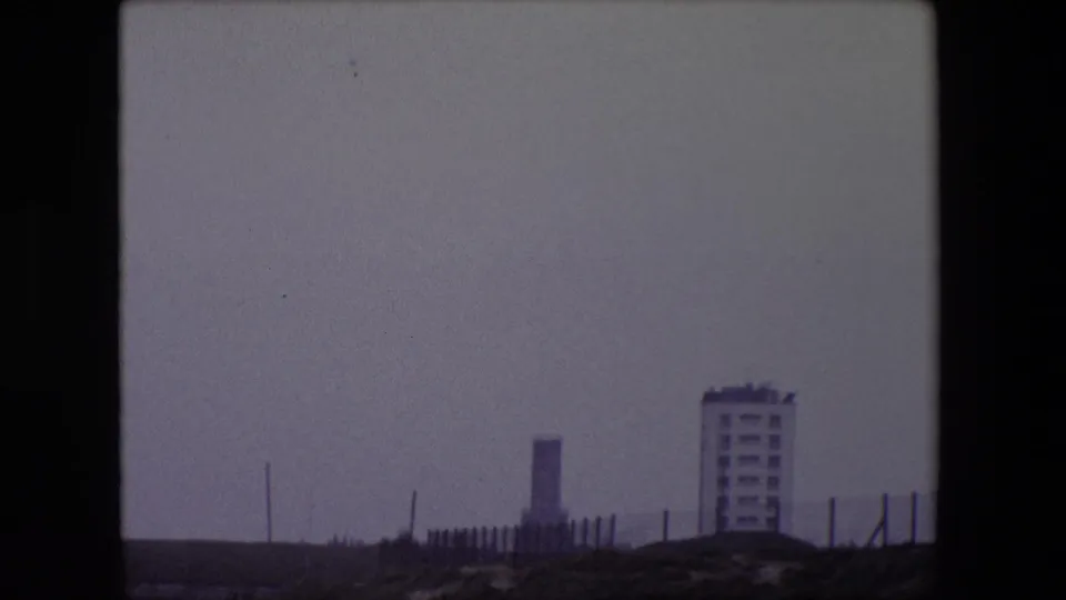 End frame of the 1967 Calais panorama showing a dark foreground embankment, fence line, vertical chimneys or masts, and a modern rectangular building against a pale sky.
