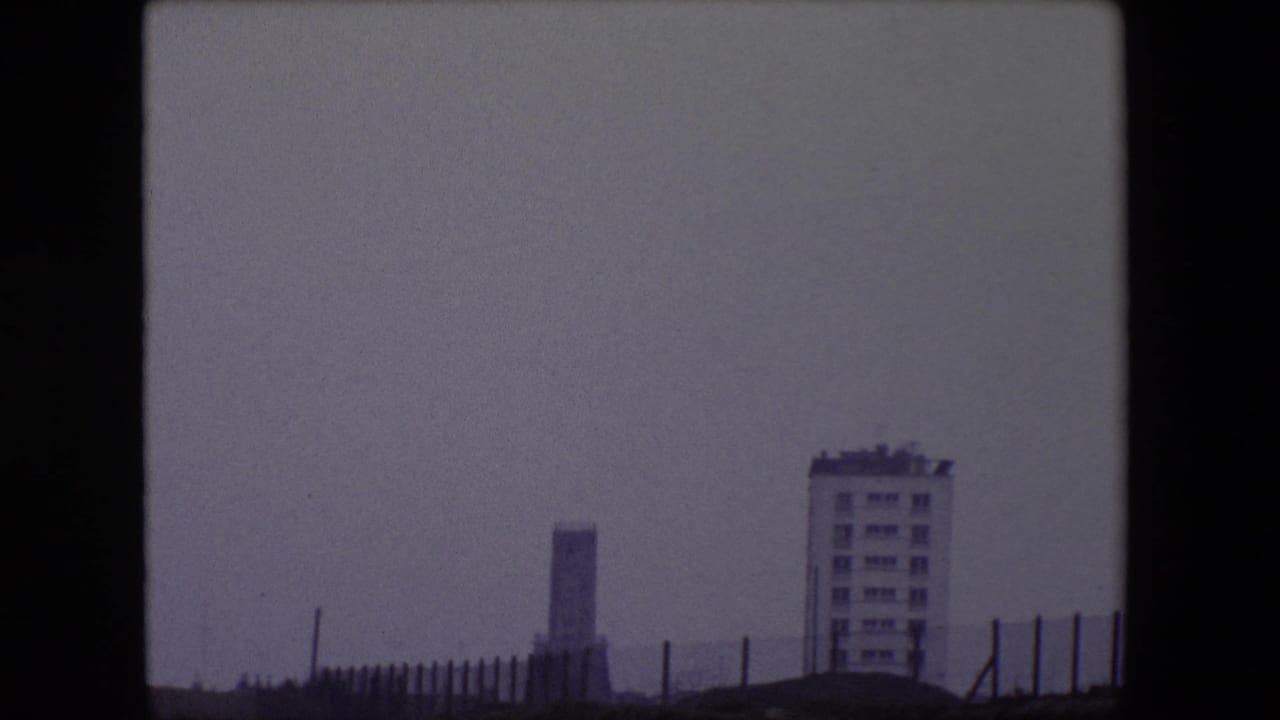 1967 Calais home-movie frame showing a hazy seafront view with a pale apartment building, a distant tower-like structure, foreground fencing, and muted gray sky.