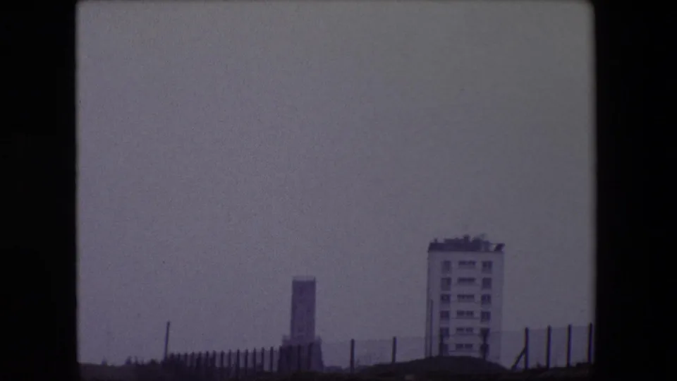 1967 Calais home-movie frame showing a hazy seafront view with a pale apartment building, a distant tower-like structure, foreground fencing, and muted gray sky.