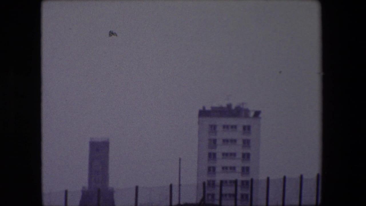 1967 Calais 8mm frame with a pale multi-story building, distant vertical structure, thin fence posts, and a hazy gray coastal horizon.