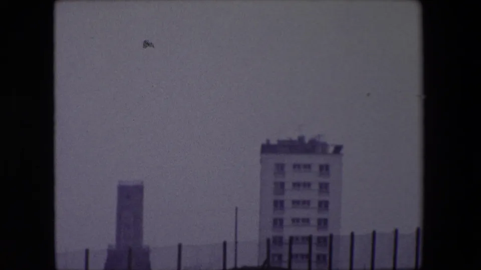 1967 Calais 8mm frame with a pale multi-story building, distant vertical structure, thin fence posts, and a hazy gray coastal horizon.