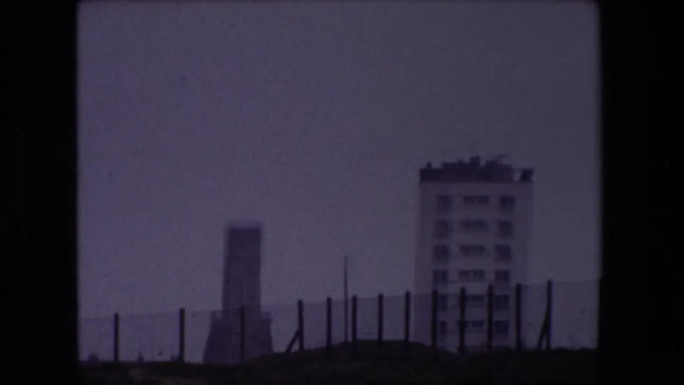 1967 Calais coastal frame showing foreground fencing, a pale apartment block, a tower-like structure near the water, and a soft gray sea horizon.