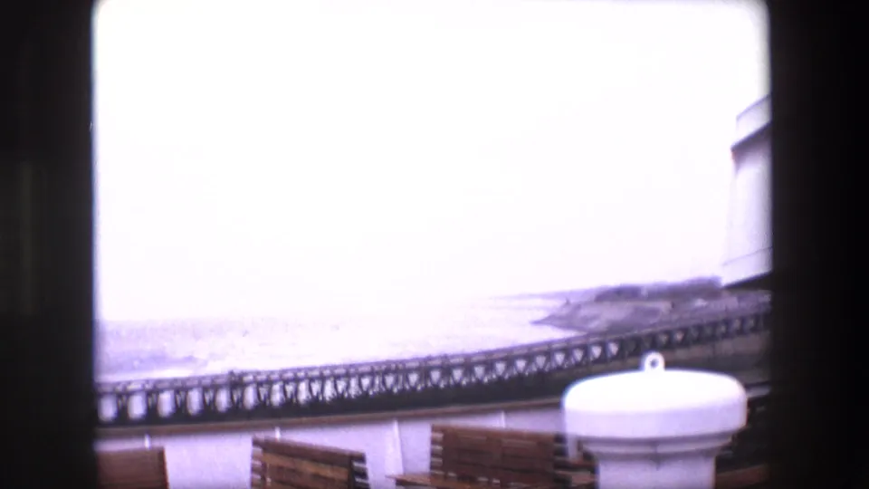 1967 Calais home-movie frame showing a bright overexposed seafront with waves, a pier or promenade railing, benches, and a white rounded coastal structure in the foreground.