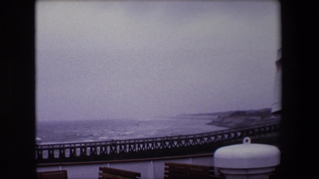 1967 8mm Calais travel frame showing a dark metal pier stretching along the coast beyond ship-deck benches and a white cylindrical fitting under a gray sky.