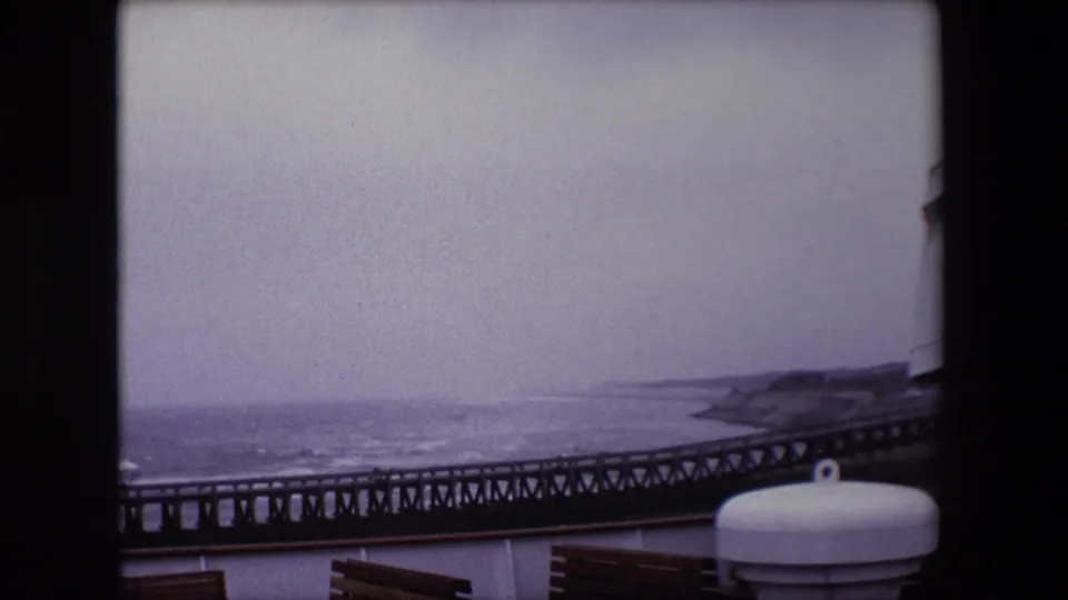 1967 8mm Calais travel frame showing a dark metal pier stretching along the coast beyond ship-deck benches and a white cylindrical fitting under a gray sky.