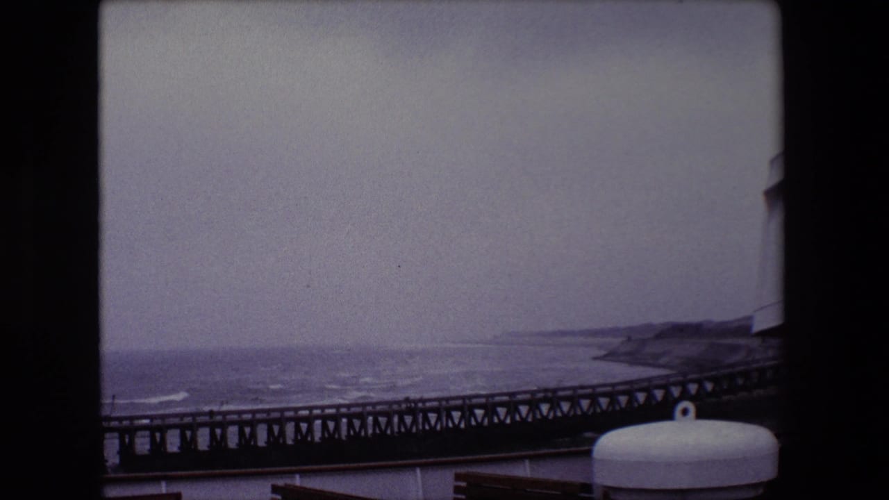 Long pier and gray sea seen from a vessel with a white deck fixture in the foreground.