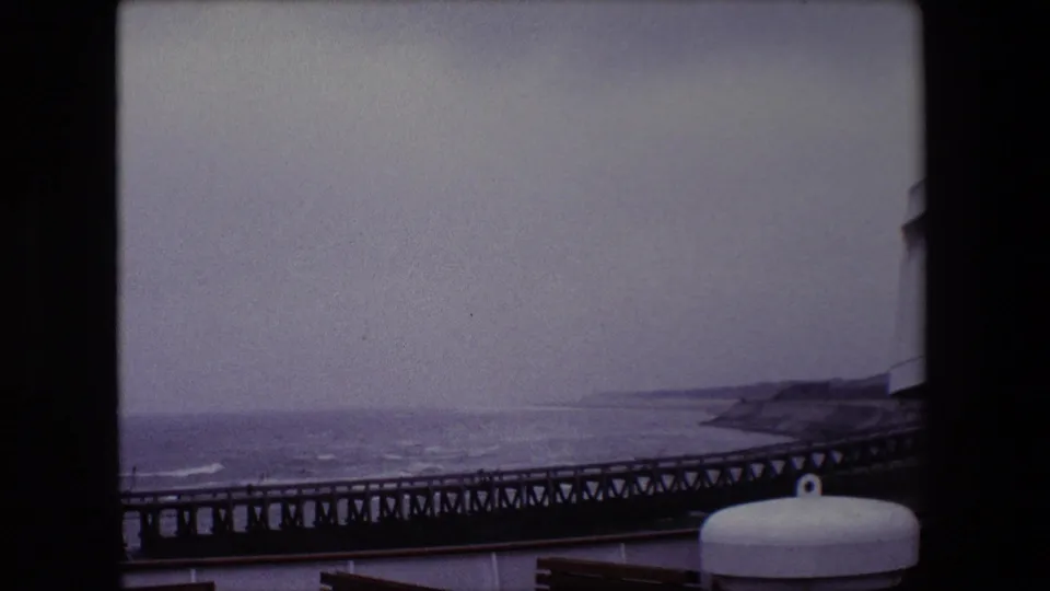 Calais, France 1967 frame from a ship deck looking past a white cylindrical deck fitting toward a long pier, rough water, and a hazy coastline.