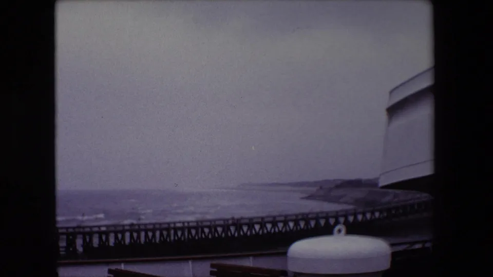 1967 8mm frame from the Calais ship-deck view with the pier low in frame, distant shoreline, wooden bench backs, a white deck fitting, and a curved ship structure at right.