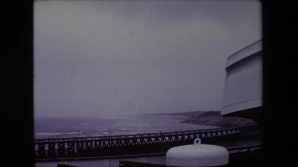 Final 1967 Calais frame looking from the vessel over a low pier line and restless water toward the coast, with a white deck fitting and curved ship structure in the foreground.