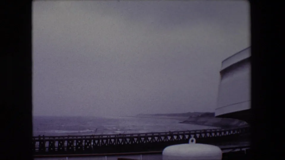 1967 Calais coastal home-movie frame showing rough water beyond a dark pier, a white cylindrical ship or pier fitting, distant shoreline, and a cropped structure at right.