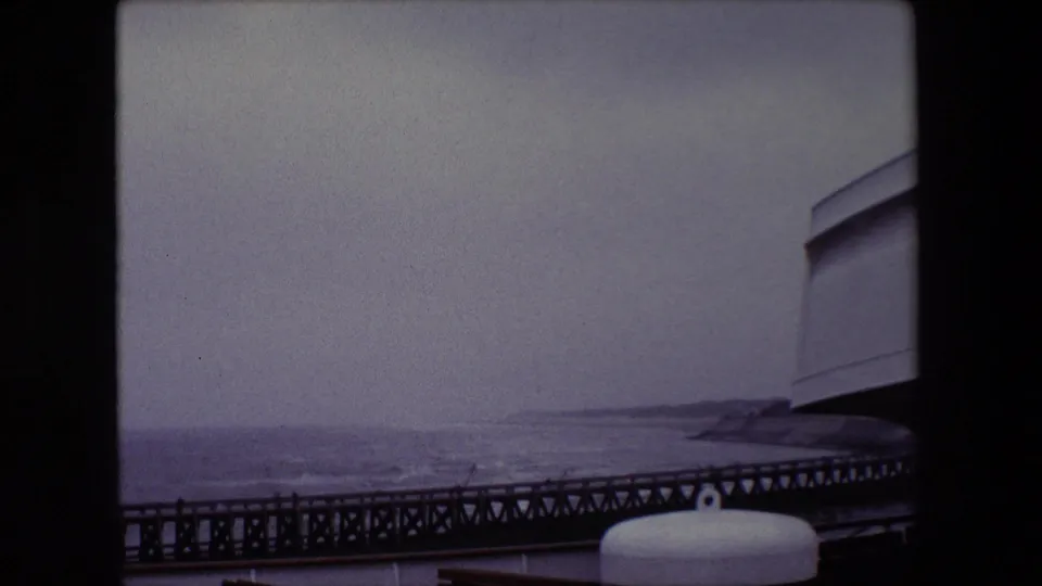 1967 Calais frame with a long pier crossing the lower view, gray Channel water, a low distant coast, and ship or pier hardware in the foreground.