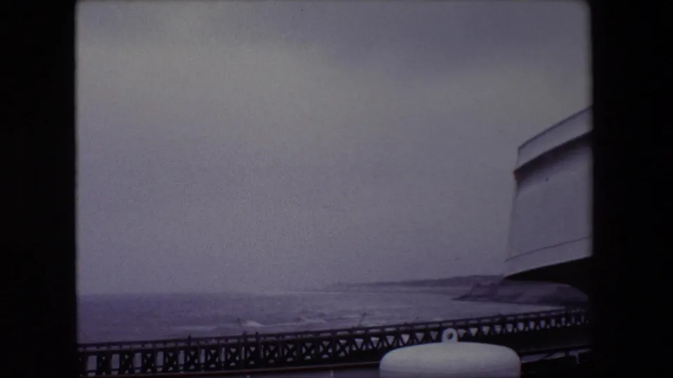 1967 Calais coastal view with softened exposure, a dark pier rail along the bottom, hazy water, and a cropped white cylindrical fitting in the foreground.
