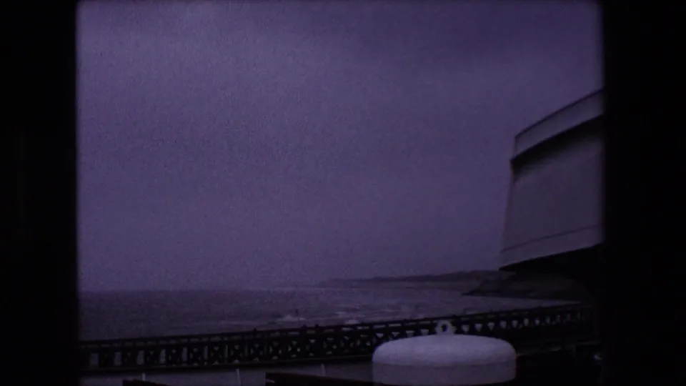 1967 Calais home-movie frame with purple-gray film color, rough water behind a dark pier, a low shoreline, and a white cylindrical object near the lower edge.
